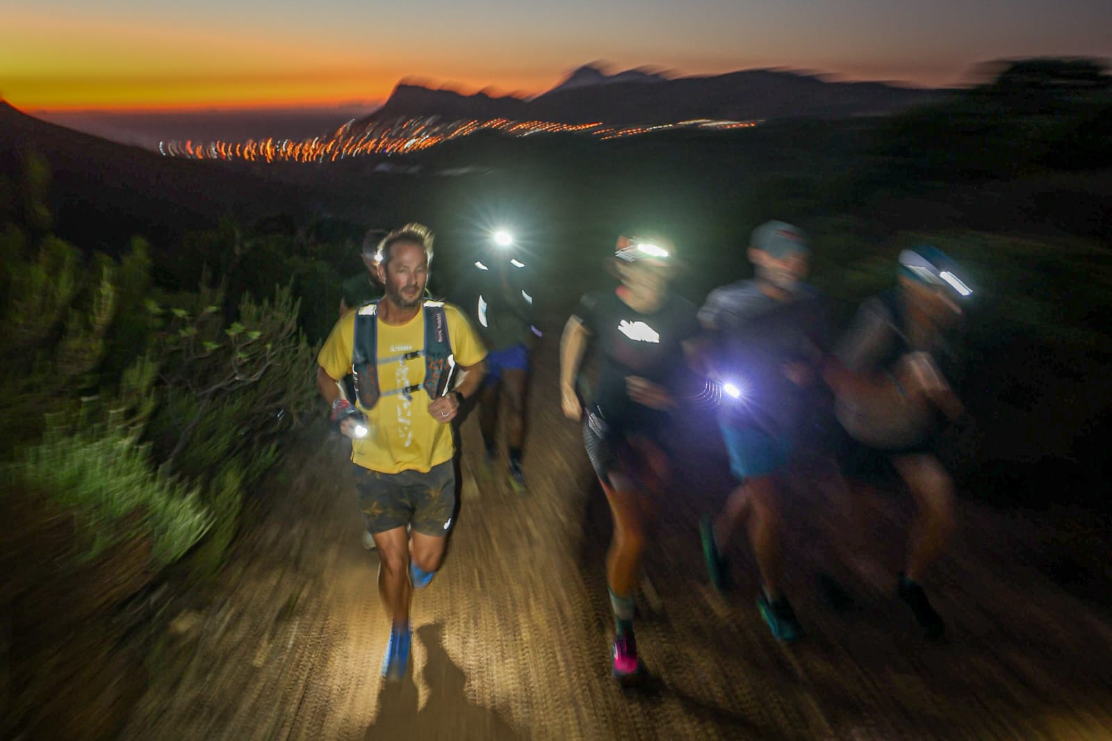 Night trail runners with headlamps against dramatic sunset backdrop with mountains and city lights